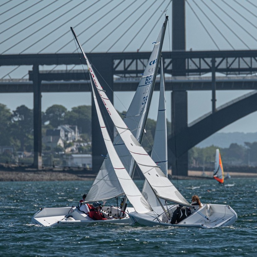 Un bateau à voile sur une étendue d'eau, il fait un virage à droite. On peut voir un pont en arrière-plant.